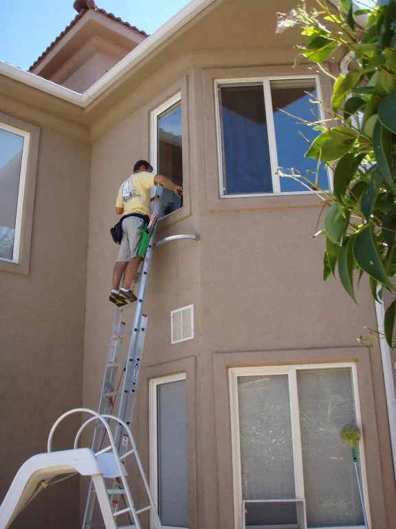 Jim cleaning an upstairs window from a ladder beside a stucco home